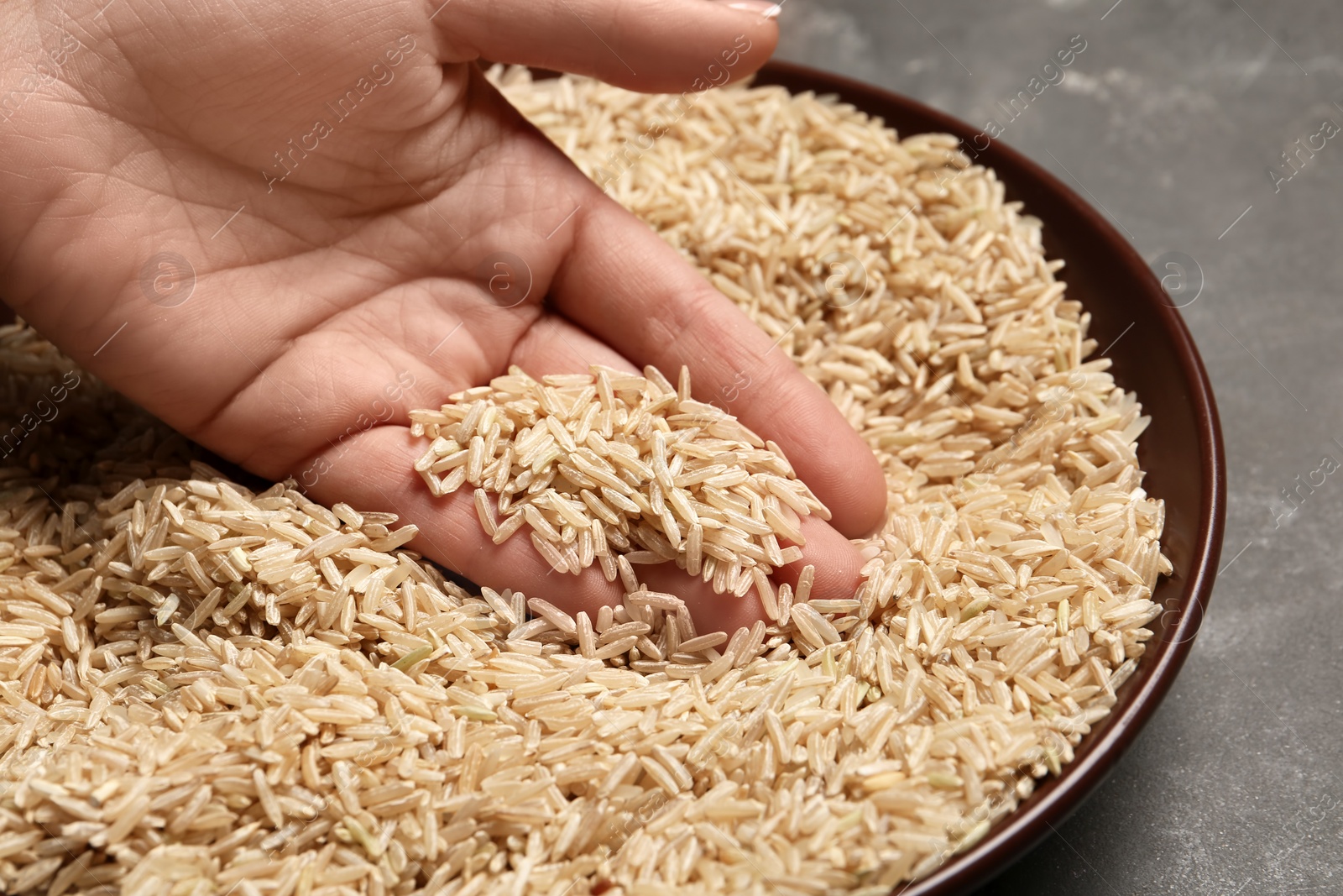 Photo of Woman holding grains near plate with brown rice on table, closeup