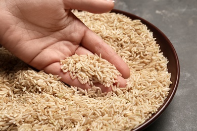 Woman holding grains near plate with brown rice on table, closeup