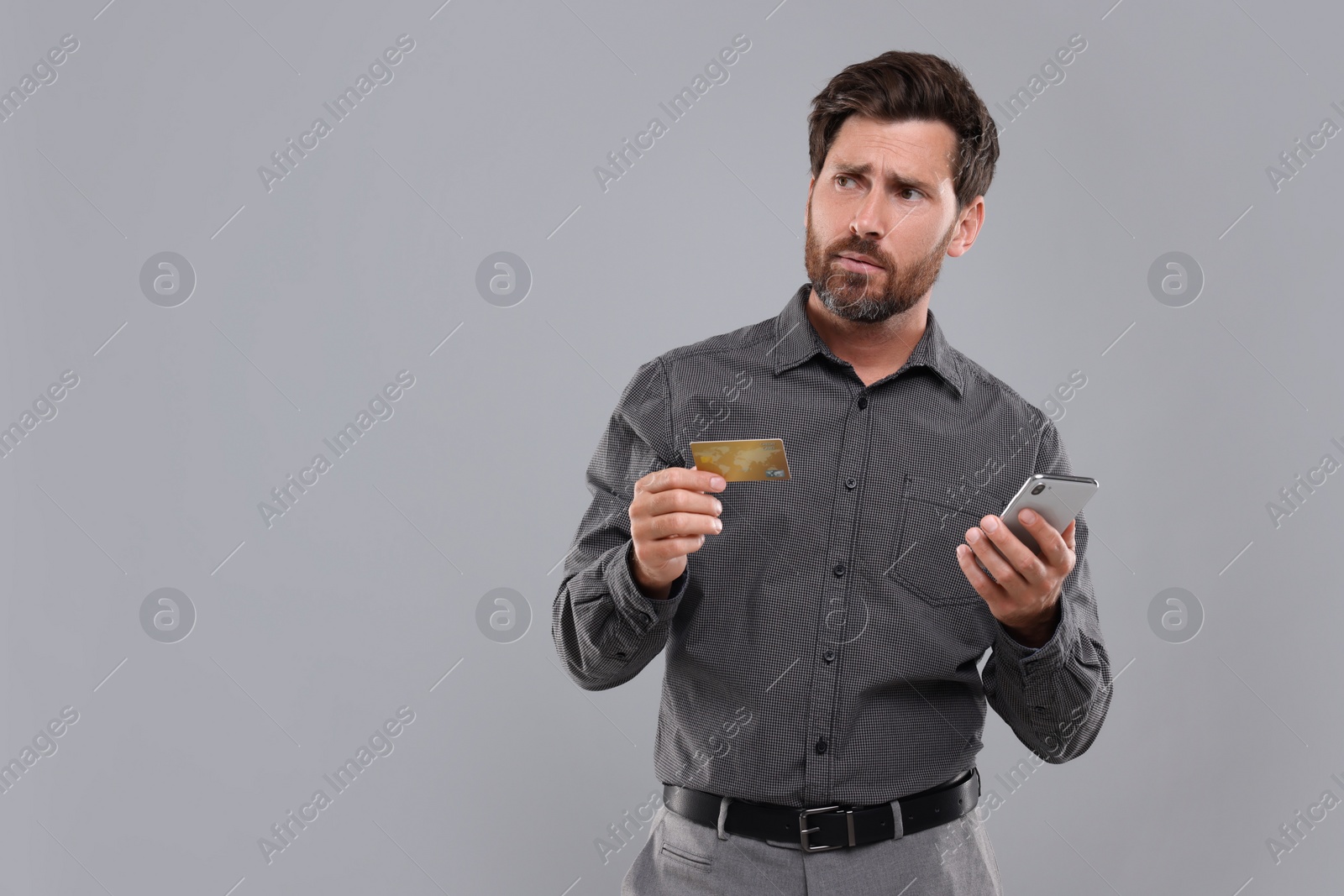 Photo of Emotional man with smartphone and credit card on light grey background, space for text. Be careful - fraud