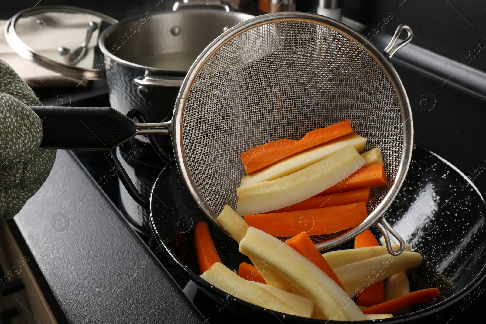 Photo of Woman putting cut parsnips and carrots into wok pan in kitchen, closeup