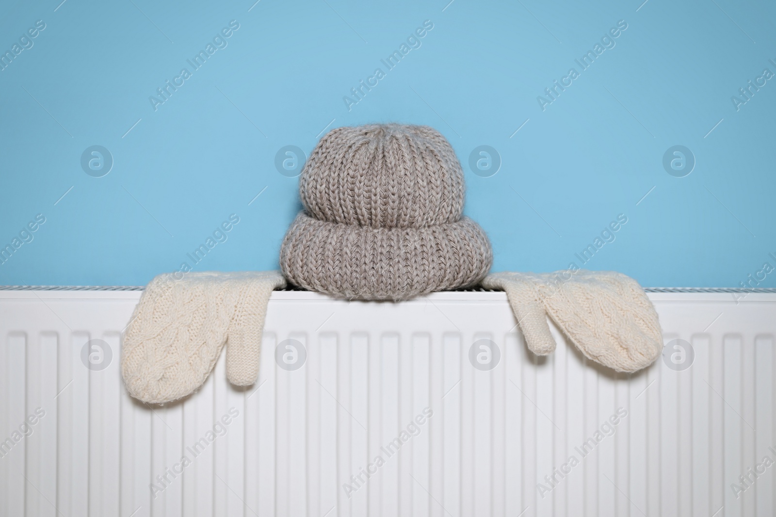 Photo of Modern radiator with knitted hat and mittens near light blue wall indoors