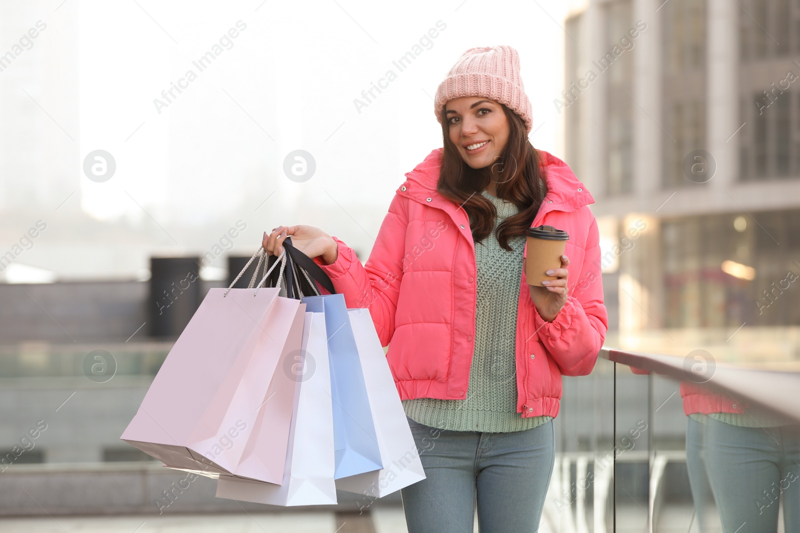 Photo of Beautiful young woman with cup of coffee and shopping bags near building