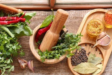 Photo of Mortar with pestle, fresh green herbs and different spices on wooden table, flat lay