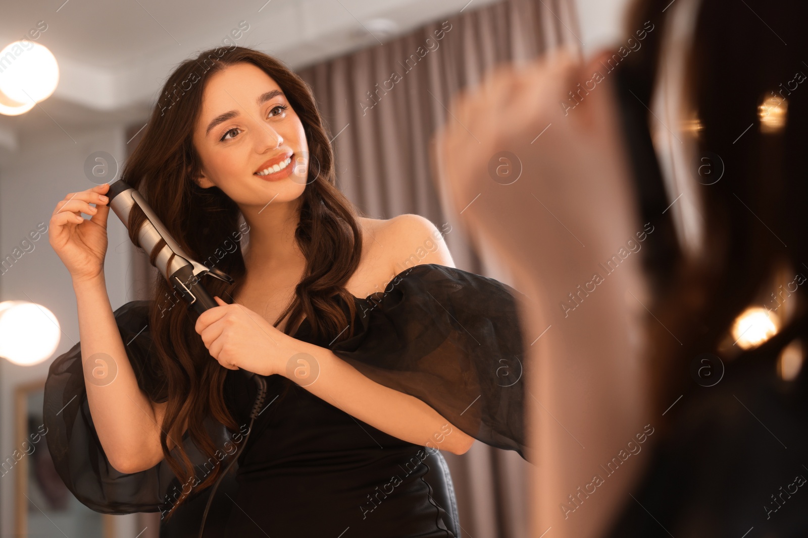 Photo of Smiling woman using curling hair iron near mirror at home