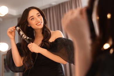 Smiling woman using curling hair iron near mirror at home