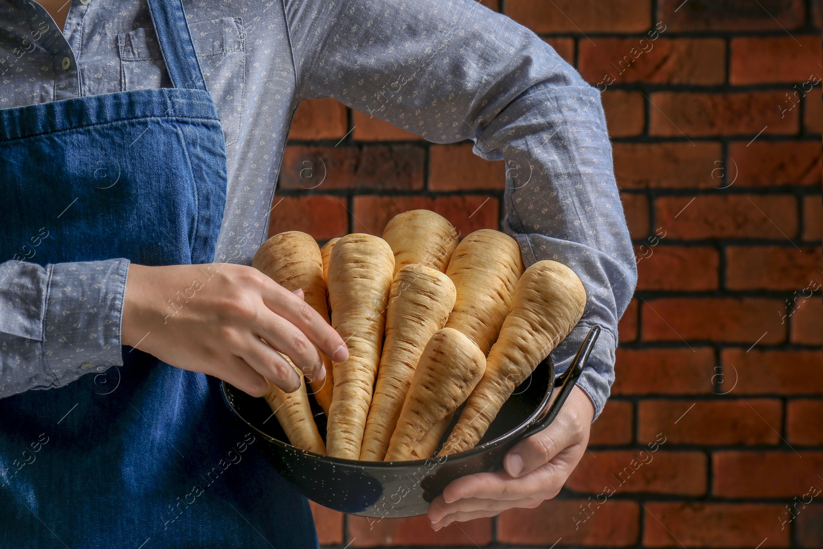 Photo of Woman holding bowl with fresh ripe parsnips near red brick wall, closeup