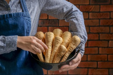 Woman holding bowl with fresh ripe parsnips near red brick wall, closeup