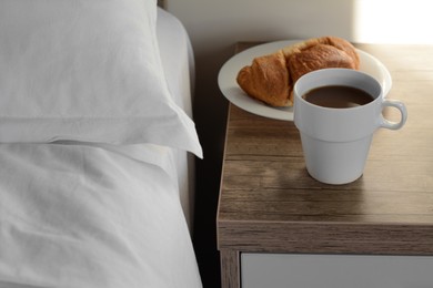 Cup of morning coffee and croissant on wooden night stand near bed indoors