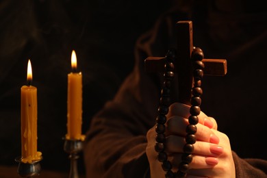 Photo of Woman with cross, beads and burning candles praying on blurred background, closeup