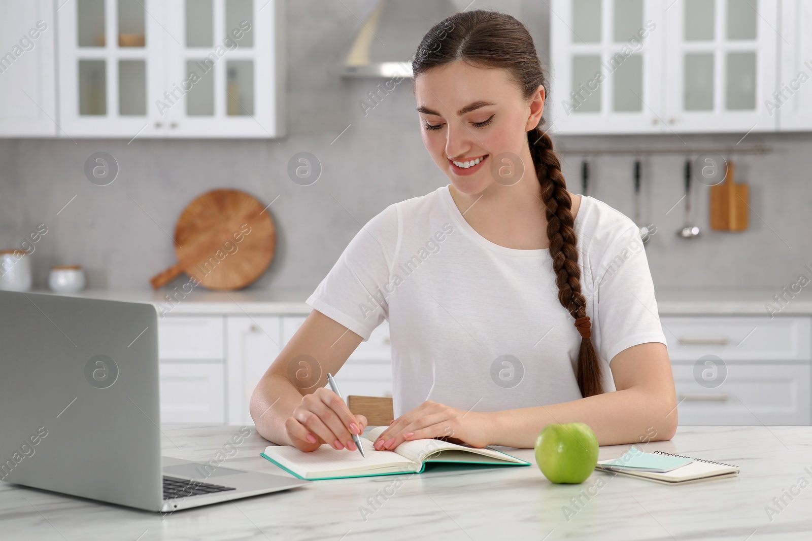 Photo of Beautiful young woman writing in notebook while working on laptop at white marble table indoors