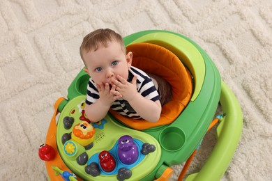 Photo of Cute baby making first steps with toy walker on soft carpet, above view