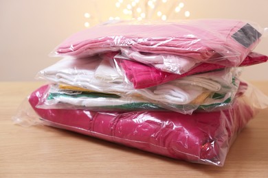 Photo of Stack of folded clothes in plastic bags on wooden table against beige background with blurred lights, closeup