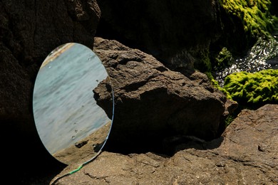 Photo of Round mirror reflecting sea on stones outdoors