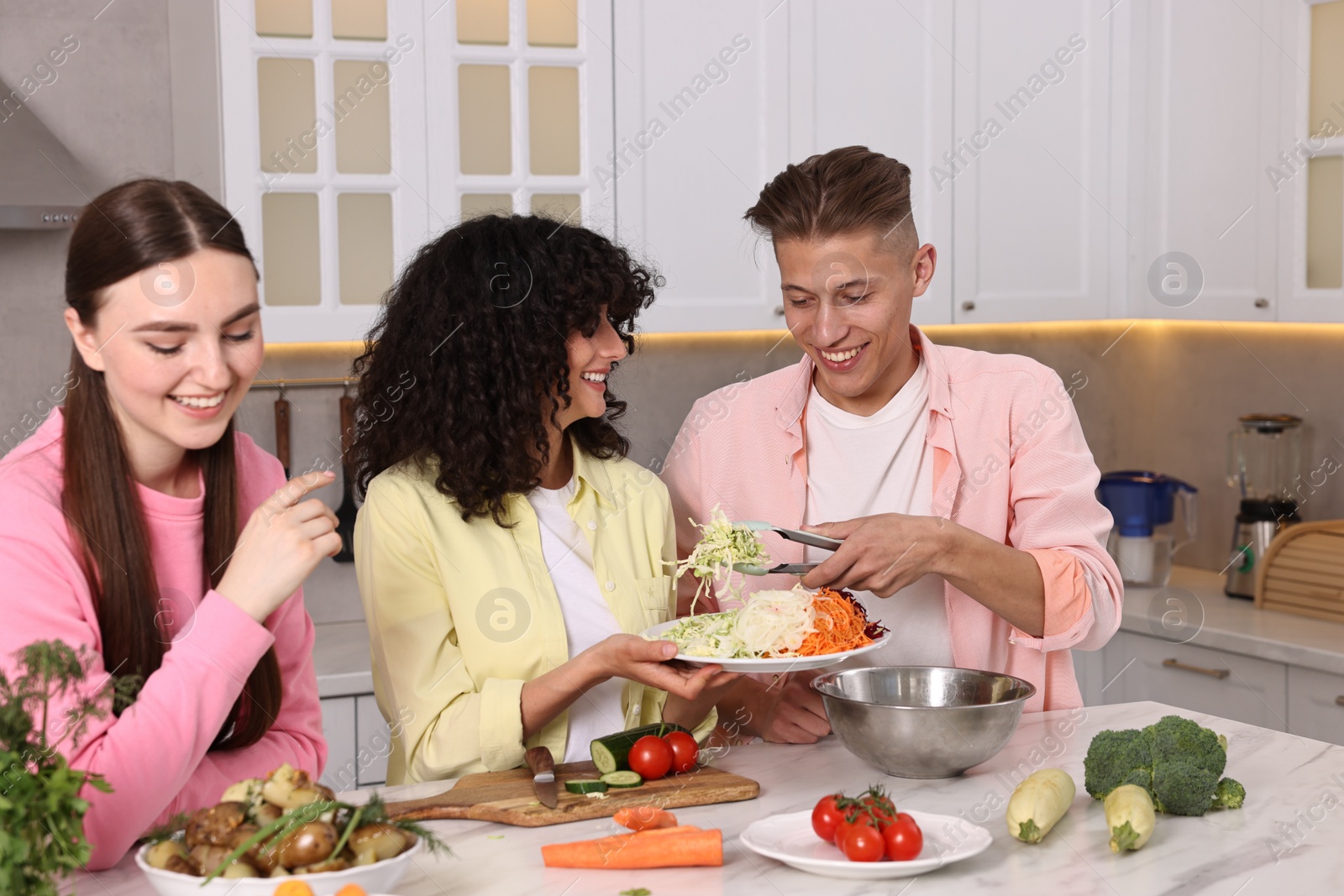 Photo of Friends cooking healthy vegetarian meal at white marble table in kitchen