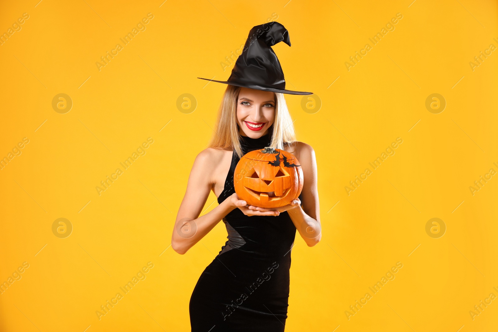 Photo of Beautiful woman in witch costume with jack o'lantern on yellow background. Halloween party