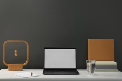 Photo of Stylish workplace with laptop, glass of water, books and decor on white table near grey wall