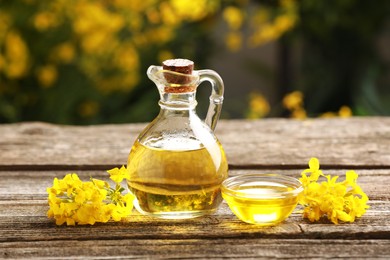 Photo of Rapeseed oil in glass jug, bowl and beautiful yellow flowers on wooden table outdoors