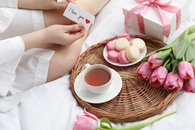 Photo of Tasty breakfast served in bed. Woman with I Love You card, macarons, tea, flowers and gift box at home, closeup