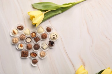 Heart made with delicious chocolate candies and beautiful tulip on beige marble table, flat lay. Space for text