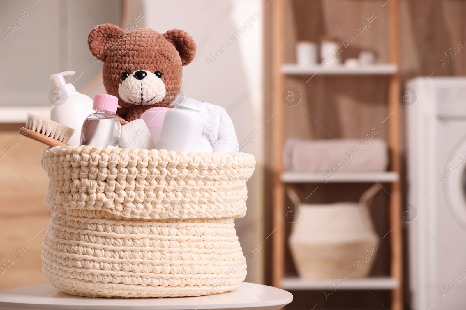 Photo of Knitted basket with baby cosmetic products, bath accessories and toy bear on white table indoors. Space for text