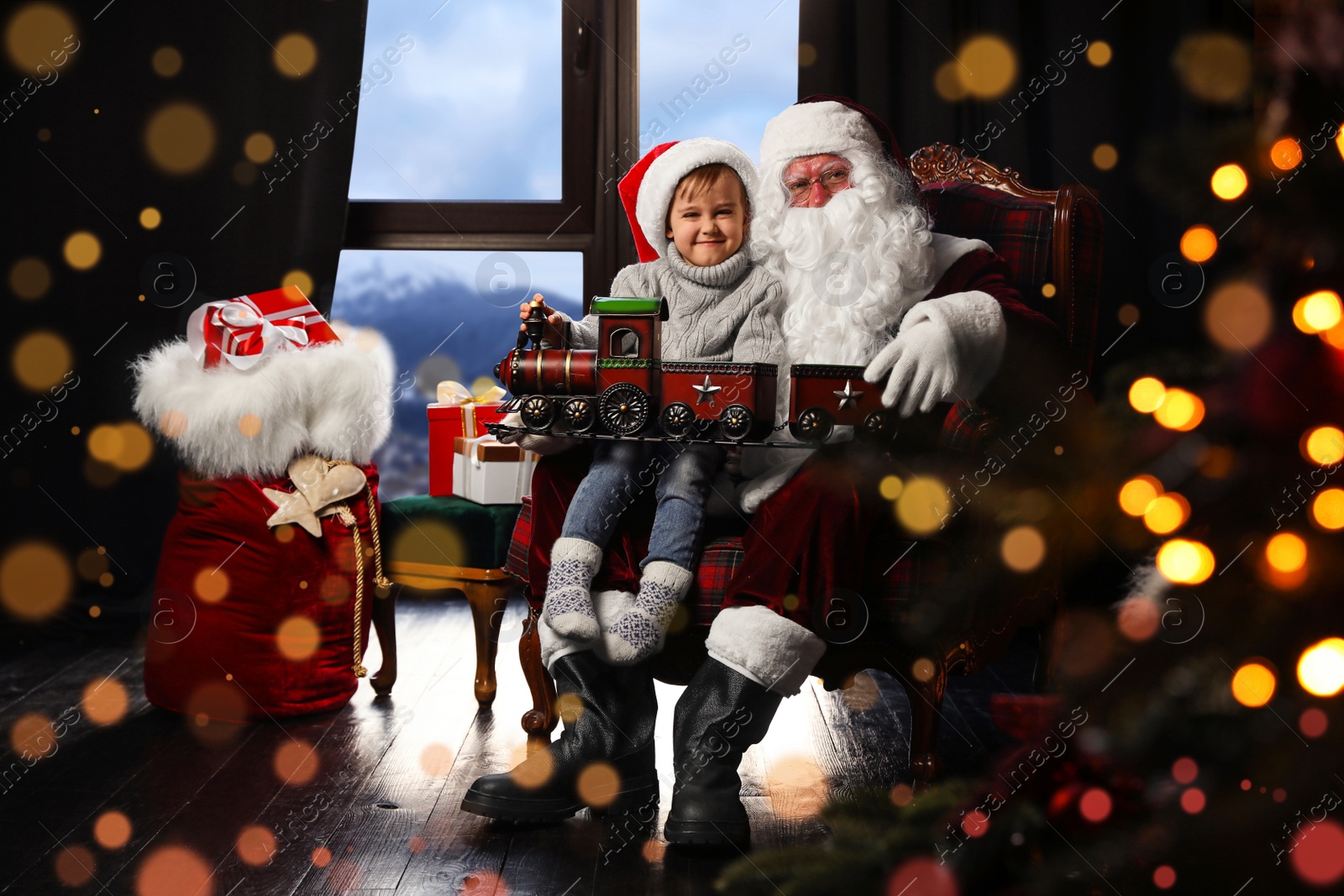 Photo of Santa Claus and little boy with toy train near window indoors. Christmas holiday