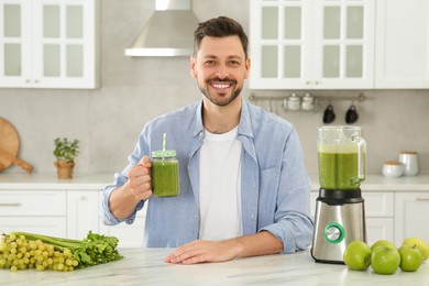 Photo of Happy man holding glass of delicious smoothie at white marble table in kitchen