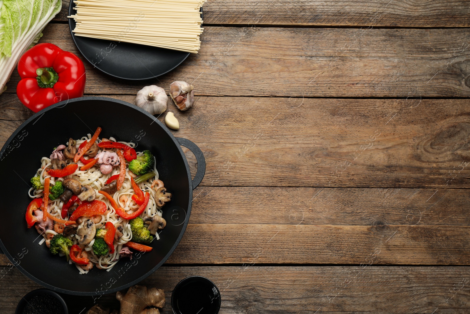 Photo of Stir fried noodles with mushrooms, seafood and vegetables in wok on wooden table, flat lay. Space for text