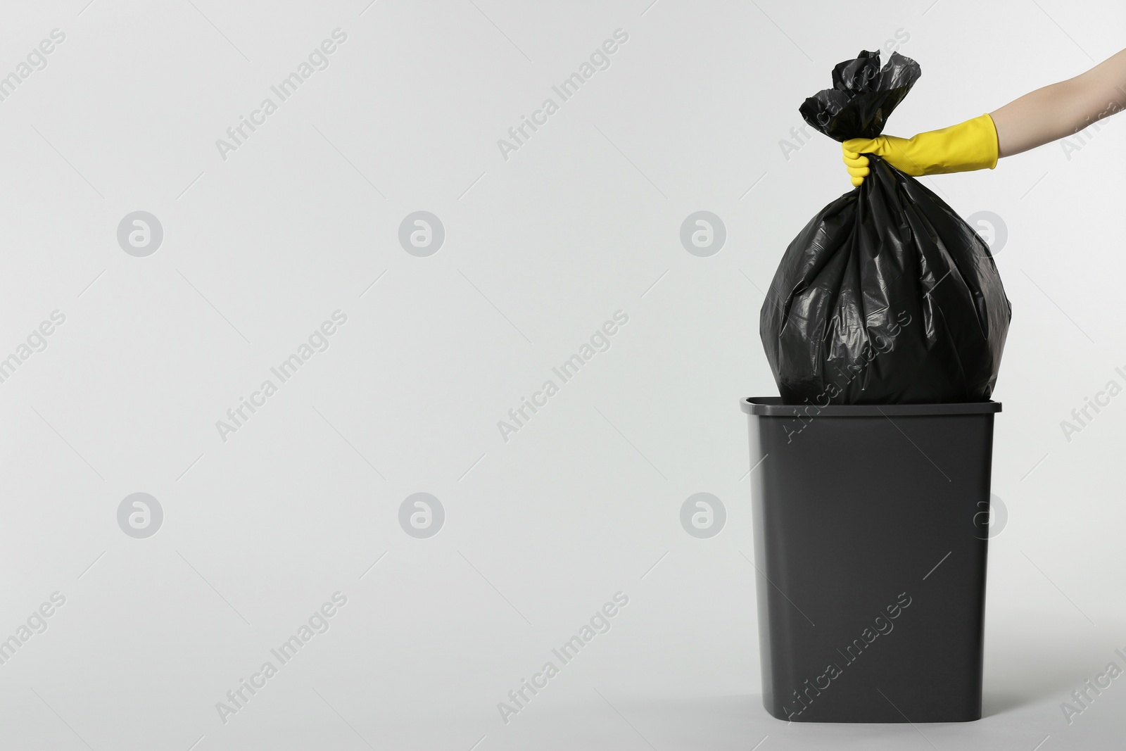 Photo of Janitor in rubber glove holding trash bag full of garbage over bucket on light grey background, closeup. Space for text