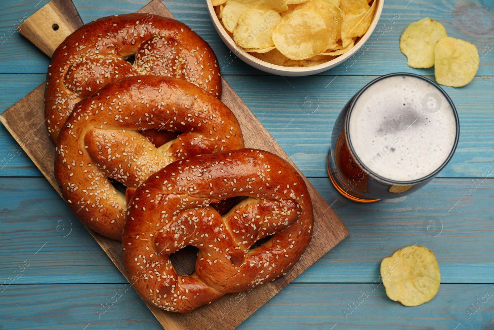 Photo of Tasty freshly baked pretzels, potato chips and glass of beer on light blue wooden table, flat lay