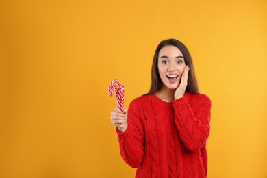 Excited young woman in red sweater holding candy canes on yellow background, space for text. Celebrating Christmas