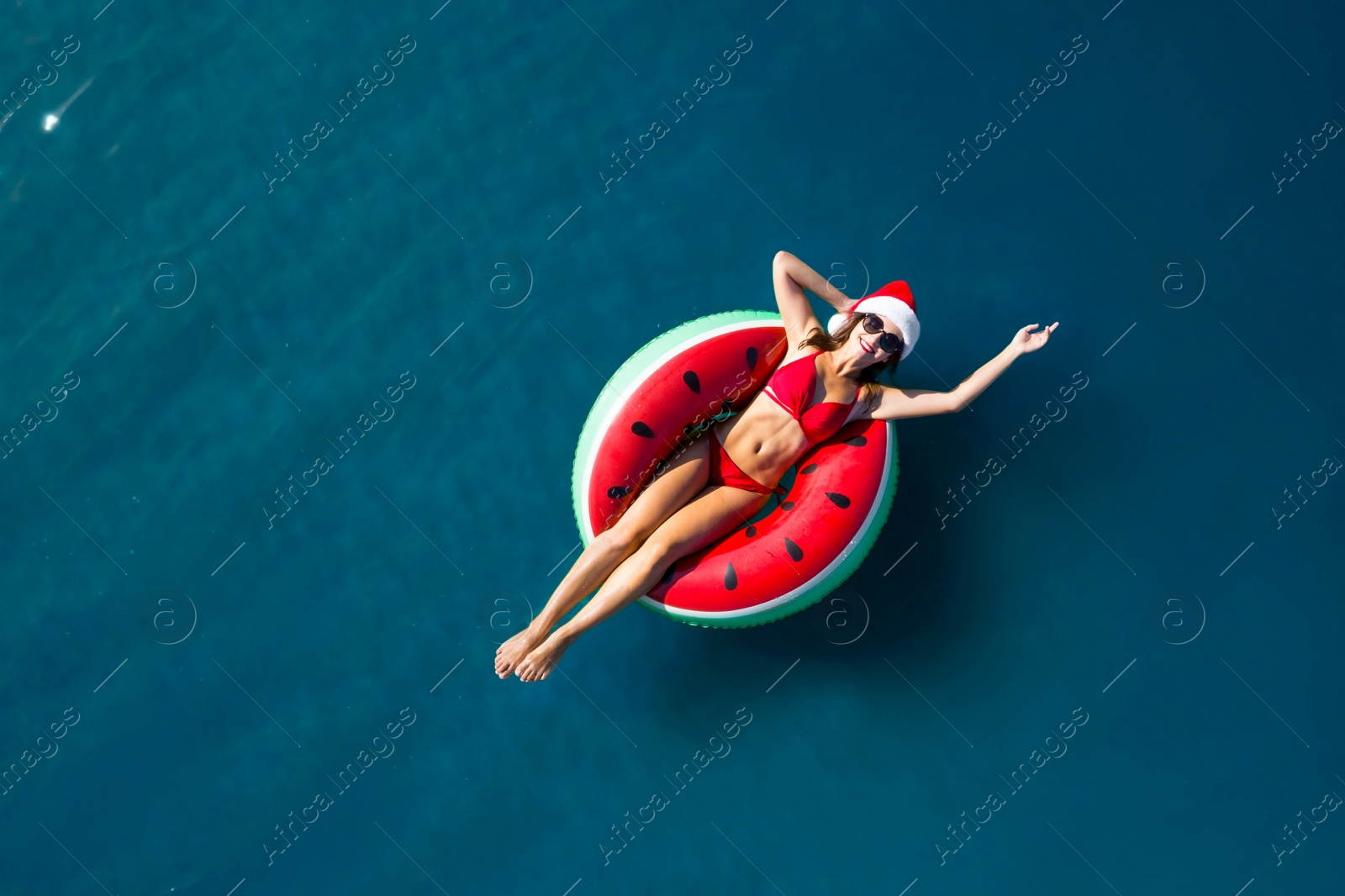 Image of Young woman wearing Santa hat and bikini with inflatable ring in sea, top view. Christmas vacation