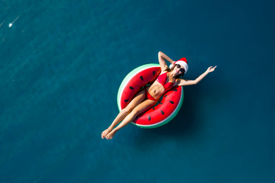 Image of Young woman wearing Santa hat and bikini with inflatable ring in sea, top view. Christmas vacation