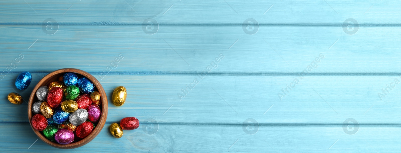 Image of Chocolate eggs wrapped in colorful foil on light blue wooden table, flat lay with space for text. Banner design