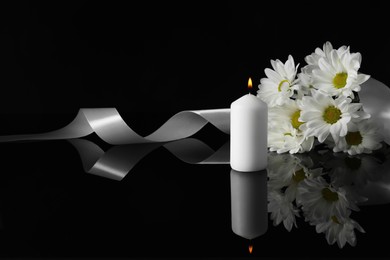 Photo of Burning candle, white chrysanthemum flowers and ribbon on black mirror surface in darkness, space for text. Funeral symbols