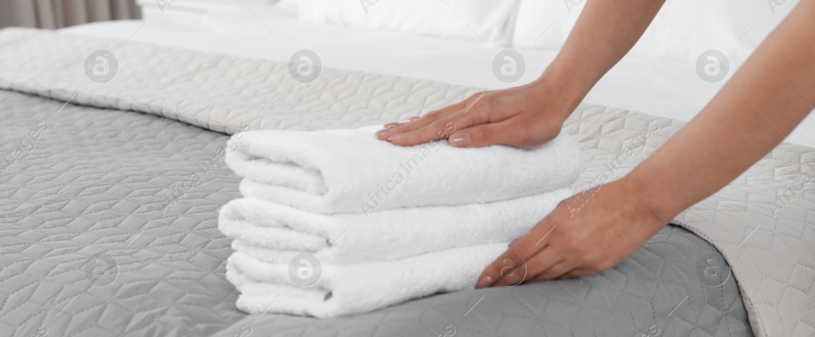 Image of Young maid putting stack of fresh towels on bed in hotel room, closeup. Banner design