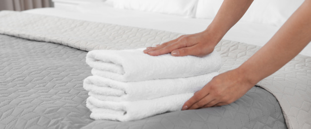 Young maid putting stack of fresh towels on bed in hotel room, closeup. Banner design