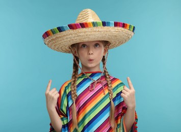 Cute girl in Mexican sombrero hat and poncho pointing at something on light blue background