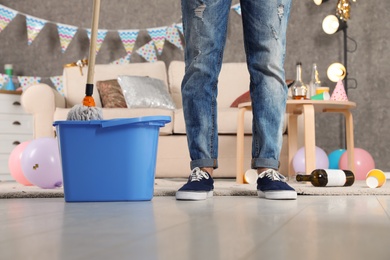 Photo of Man with mop and bucket cleaning messy room after party, closeup view of legs