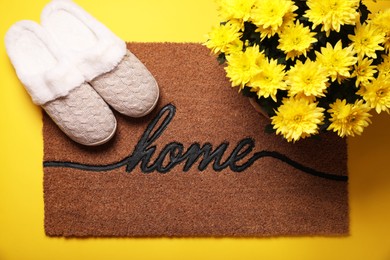 Photo of Doormat with word Home, slippers and flowers on yellow background, flat lay