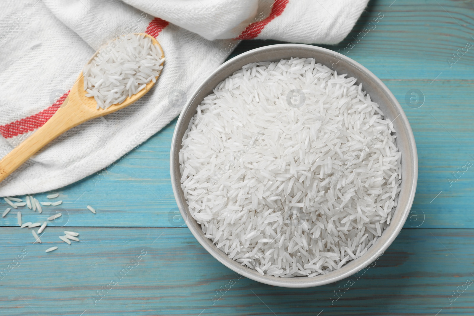 Photo of Raw basmati rice in bowl and spoon on light blue wooden table, flat lay