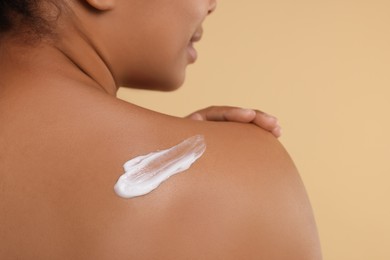 Young woman applying body cream onto back on beige background, closeup