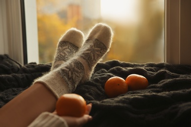 Woman in knitted socks and tangerines on plaid near window at home, closeup