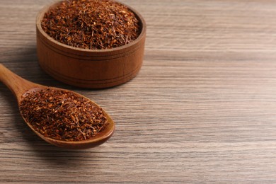 Spoon and bowl with dry rooibos leaves on wooden table, space for text