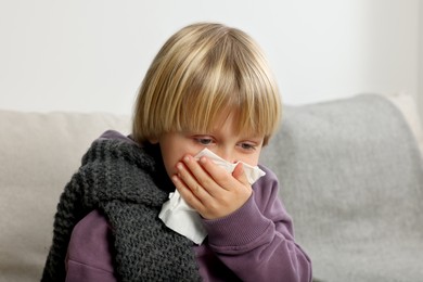 Photo of Boy blowing nose in tissue on sofa in room. Cold symptoms