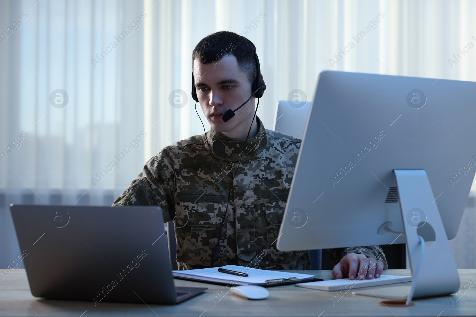 Photo of Military service. Young soldier in headphones working at wooden table in office