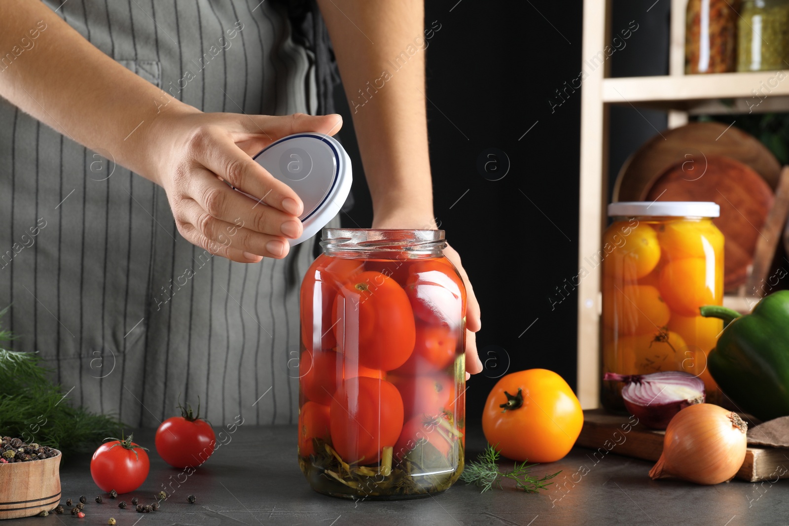 Photo of Woman closing jar with pickled tomatoes at grey table, closeup