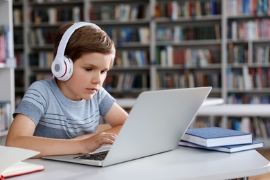 Photo of Little boy with headphones reading book using laptop in library