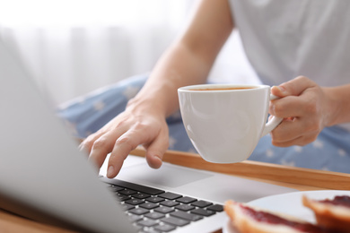 Woman with cup of morning coffee working on laptop indoors, closeup