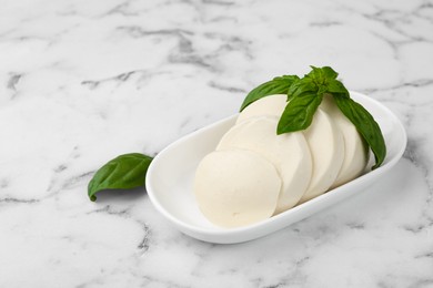 Plate with tasty mozzarella slices and basil leaves on white marble table. Space for text