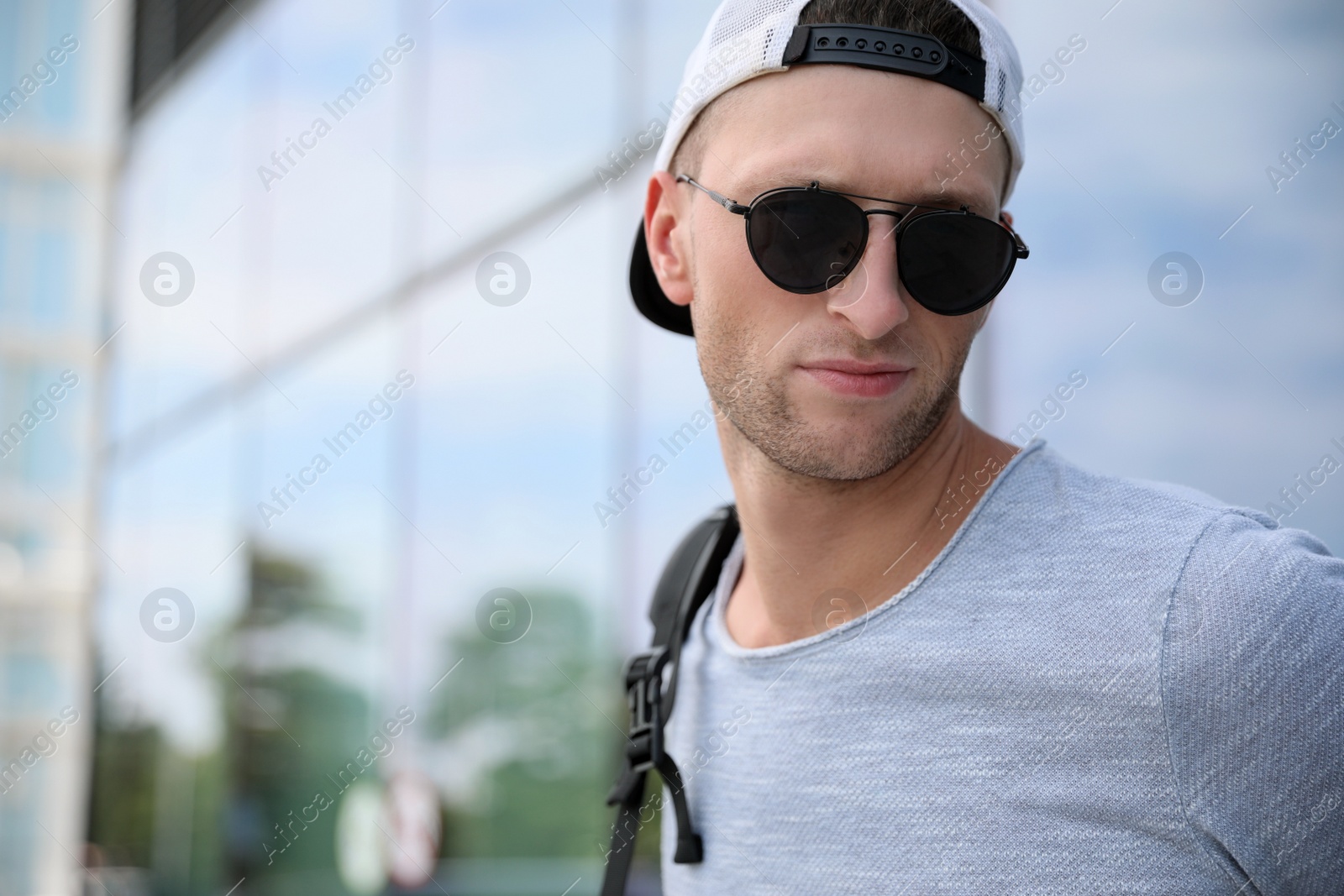 Photo of Handsome young man with stylish sunglasses and backpack near reflection surface outdoors, space for text